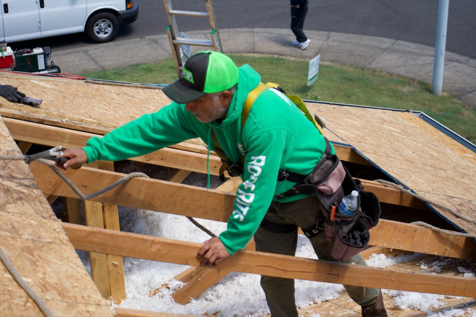 Man in green working on a roof in Salem OR, wearing a harness, with a van in the background.