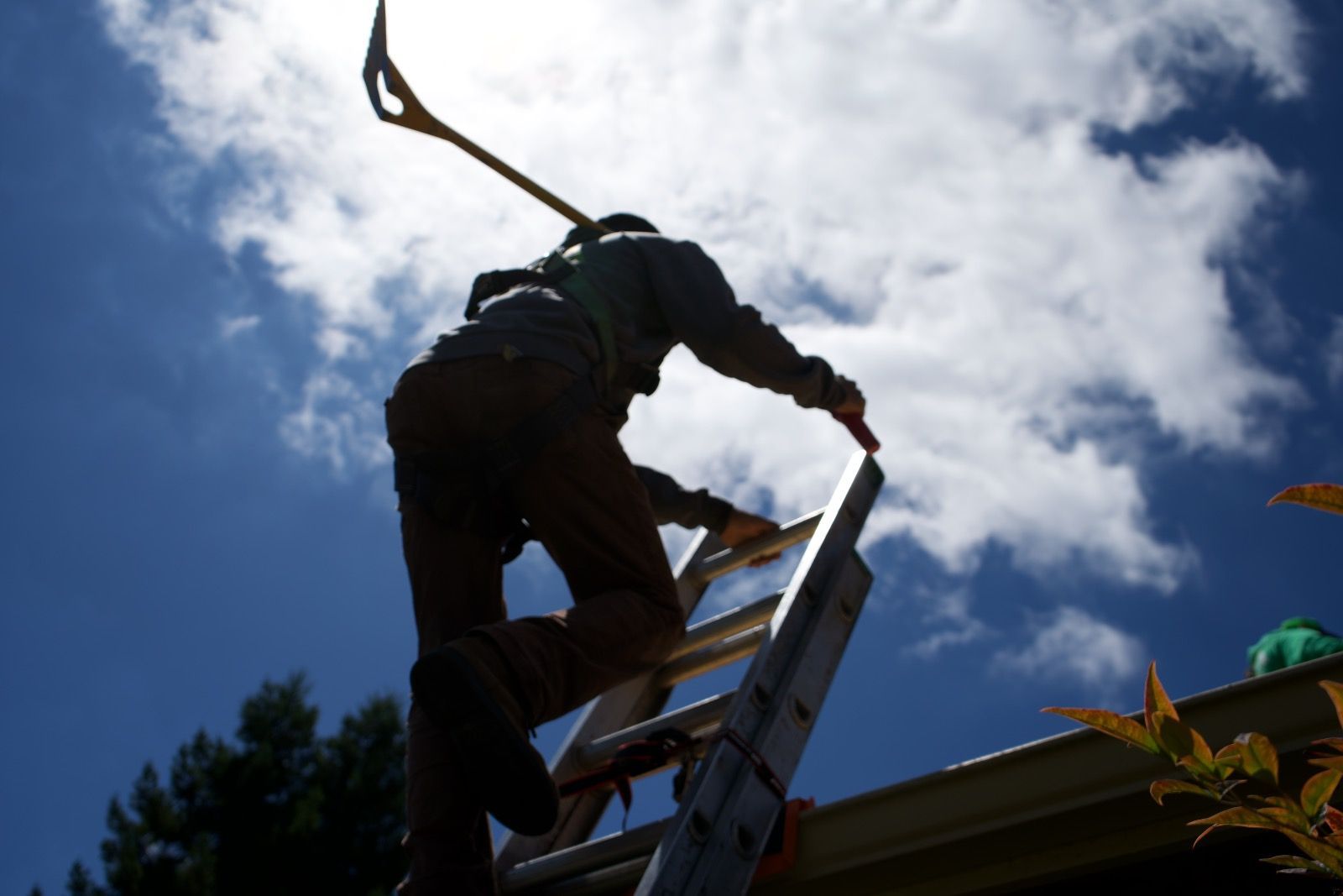 Person climbing a ladder to a roof, holding a shovel, bright sun and blue sky.
